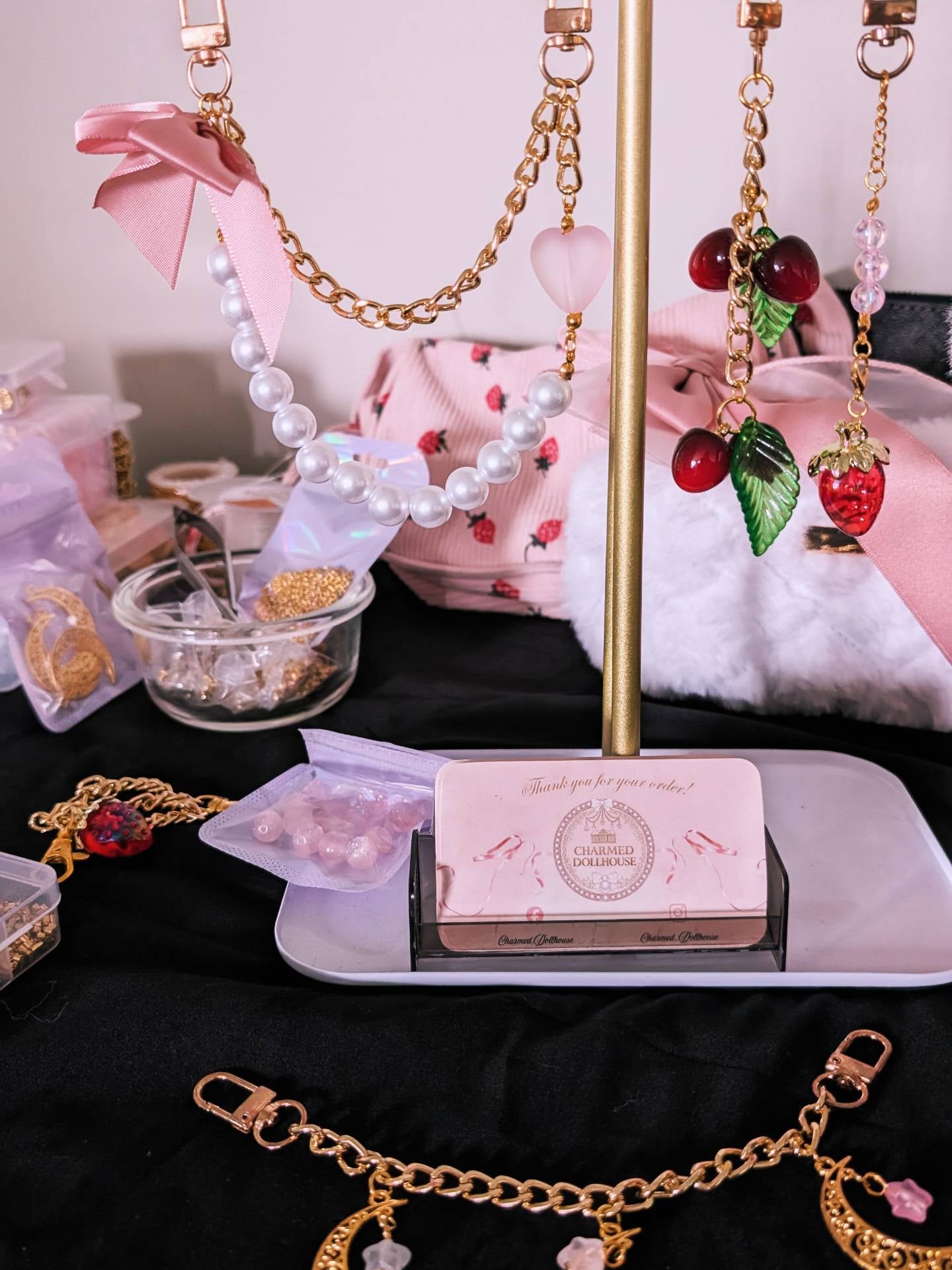 A styled jewelry display featuring gold-tone chain necklaces, pearl strands, strawberry-themed earrings, and a pink box labeled 'Cherry Blossom' on a stand over a black cloth.