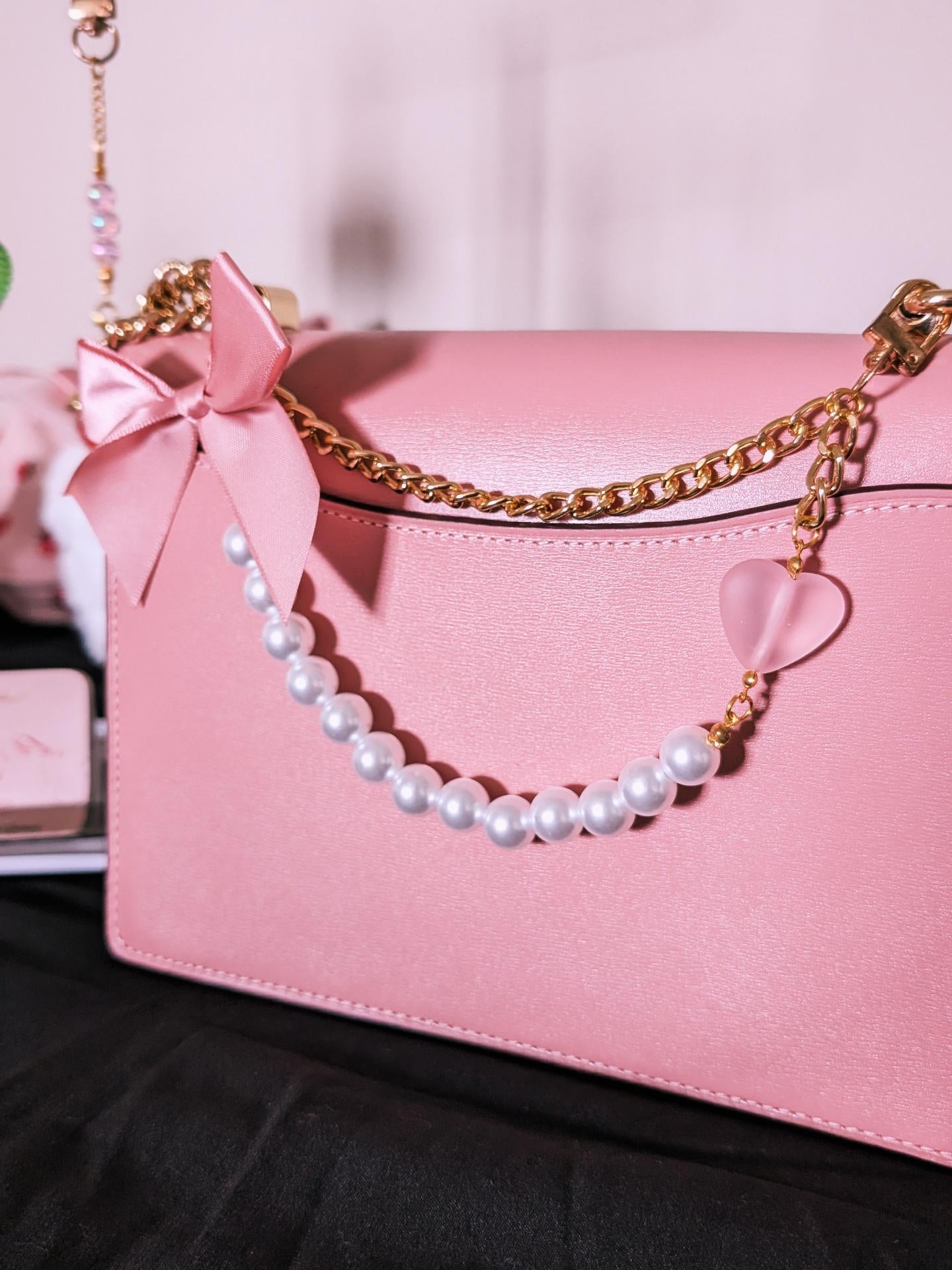 A pink leather shoulder bag with a gold chain strap decorated with white faux pearls, a pink satin bow, and a translucent pink heart charm, resting on a black surface.