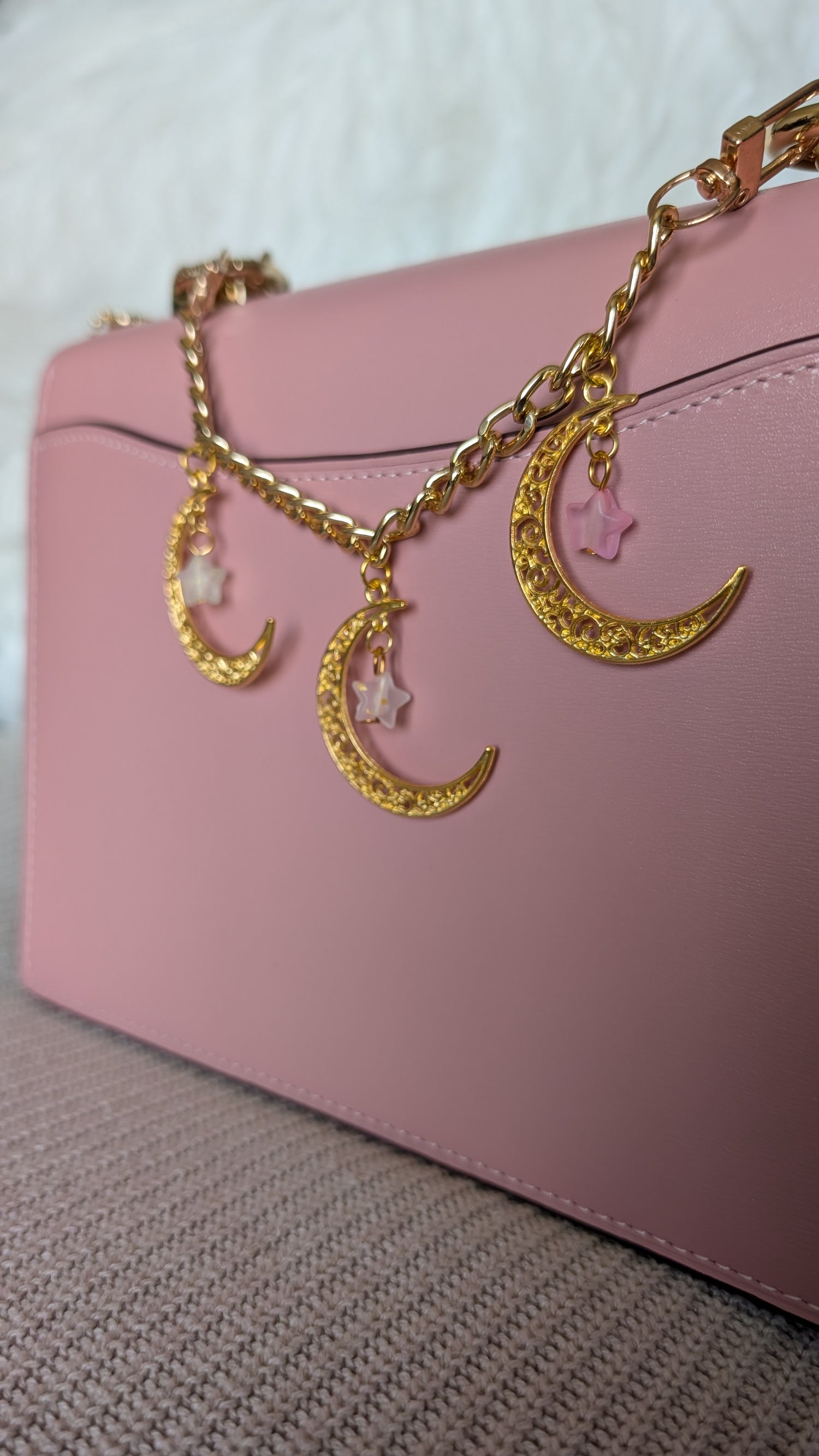 Pink handbag with gold chain and moon and star charms on a textured surface.